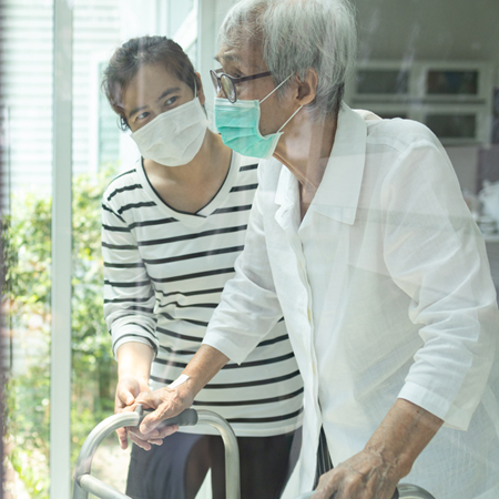 Frau begleitet Seniorin mit Gehhilfe vor Fensterfront