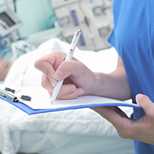 Ärztin unterschreibt ein Dokument auf einem Klemmbrett, im Hintergrund ein Patient im Krankenbett