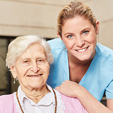 Ältere Frau im Rollstuhl, hinter ihr eine weibliche, junge Pflegekraft. Beide lächeln.