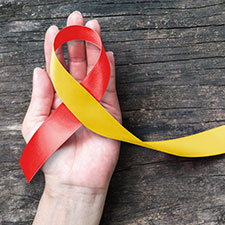 Foto: Eine offene Hand hält ein Band, das zu einer Schleife gebunden ist und auf der einen Seite rot, auf der anderen gelb ist.