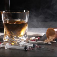 Whiskey im Glas, Spritzen, Pillen und Zigaretten auf grauem Tisch