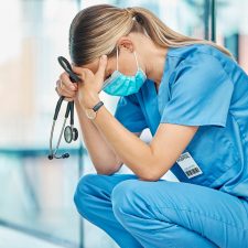 Krankenschwester kauert erschöpft mit Stethoskop in der Hand auf Krankenhausflur