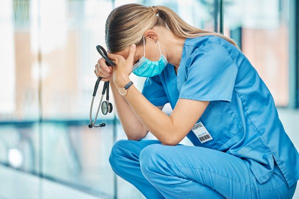 Krankenschwester kauert erschöpft mit Stethoskop in der Hand auf Krankenhausflur