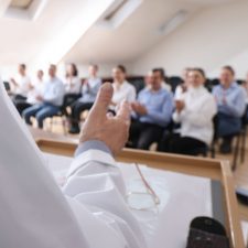 Pflegekräfte sitzen auf Stühlen in einem Seminarraum. Am Redepult steht ein Referent.