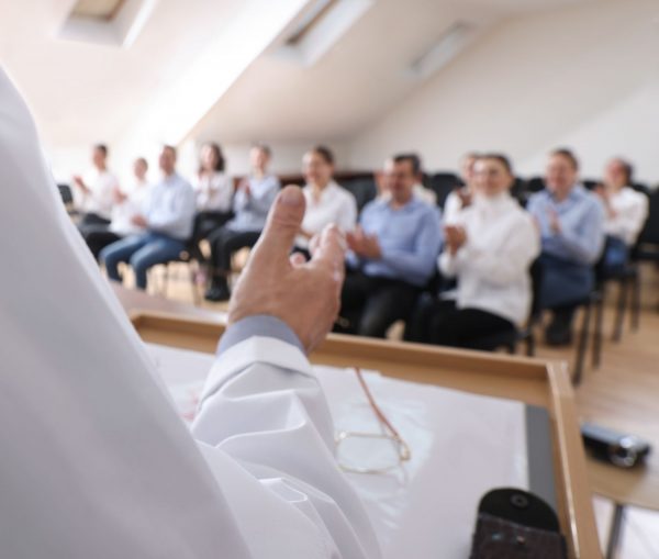 Pflegekräfte sitzen auf Stühlen in einem Seminarraum. Am Redepult steht ein Referent.