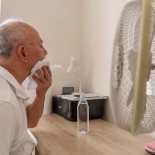 Alter Mann leidet unter Hitze und sitzt mit Ventilator und Wasserflasche am Tisch
