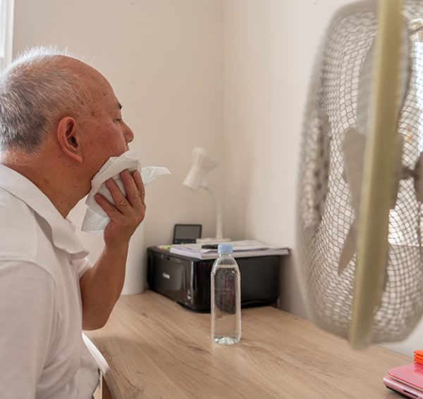 Alter Mann leidet unter Hitze und sitzt mit Ventilator und Wasserflasche am Tisch