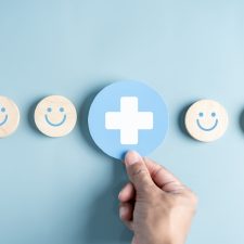 Auf blauem HIntergrund liegen lächelnde Smileys. Eine Hand legt ein blau-weißes Kreuz der Pflege ab