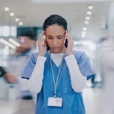Krankenschwester steht im Krankenhausflur und fasst sich mit beiden Händen an die Schläfen