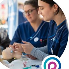 Foto aus Veranstaltungsflyer. Zwei Krankenpflegerinnen üben mit Stethoskop an einer Puppe